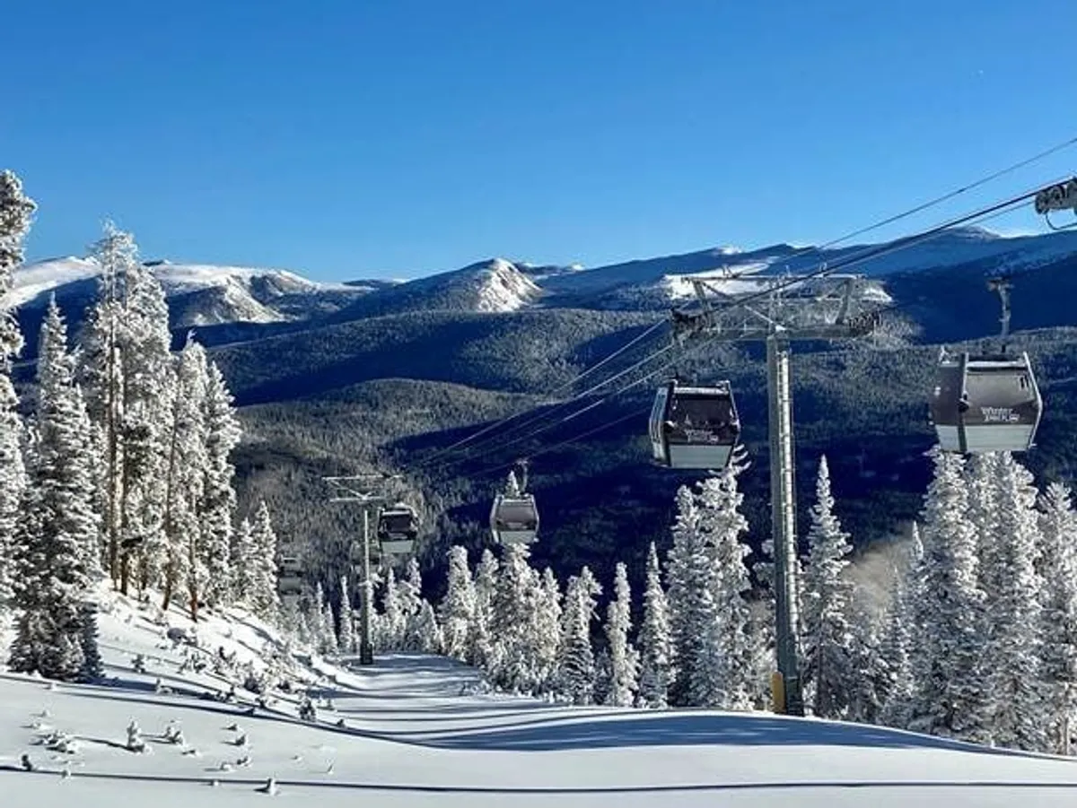 A sweeping exterior shot of a ski lift ascending beside snow-covered trees, with a majestic mountain range under a clear blue sky. The snow is pristine, and the environment exudes the thrill and natural beauty of winter sports.