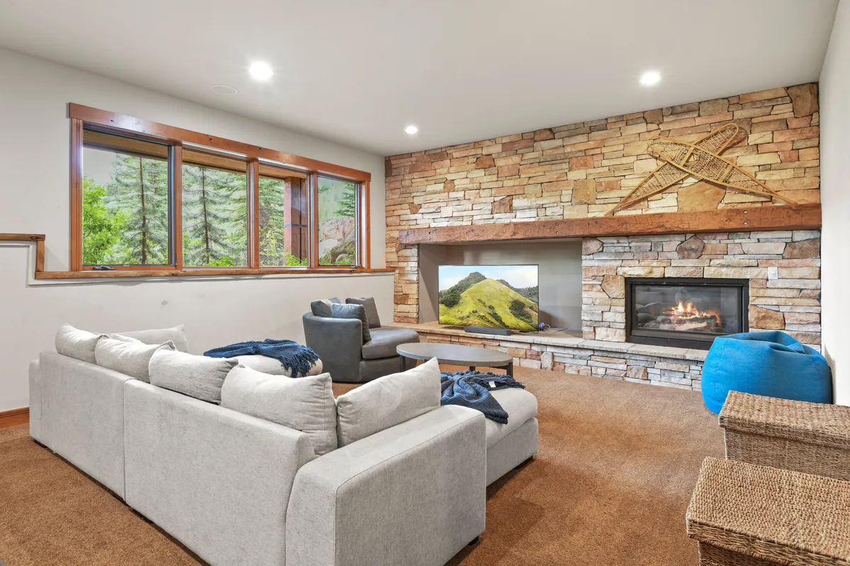 A spacious lower-level living area featuring a stone fireplace, a large sectional sofa, a TV with a landscape image, and big windows letting in natural light. A blue bean bag adds a touch of playful comfort, inviting relaxed movie nights or social gatherings.