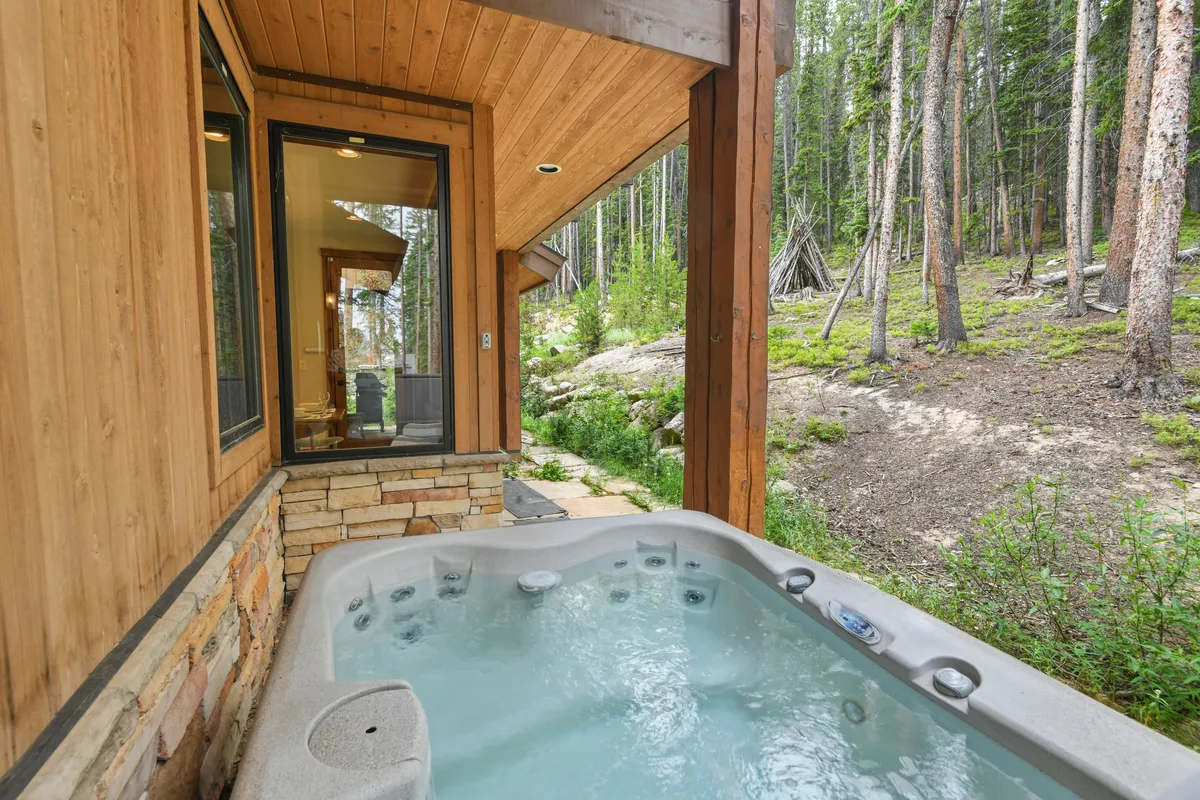 An exterior view of a luxurious wooden chalet nestled among trees, boasting a private hot tub on the deck. The setting is tranquil, with steam rising from the hot tub and nature all around, inviting relaxation after a day on the slopes.