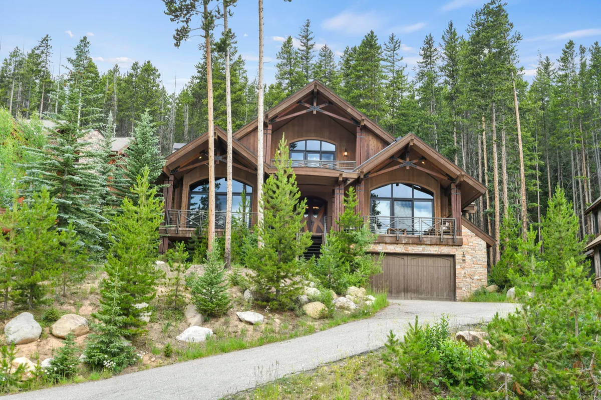The image showcases the impressive exterior of a luxury chalet nestled among tall trees, with large windows reflecting golden sunset hues. The inviting main entrance opens to a spacious balcony perfect for gathering, while the tranquil mountain setting hints at endless outdoor adventures.
