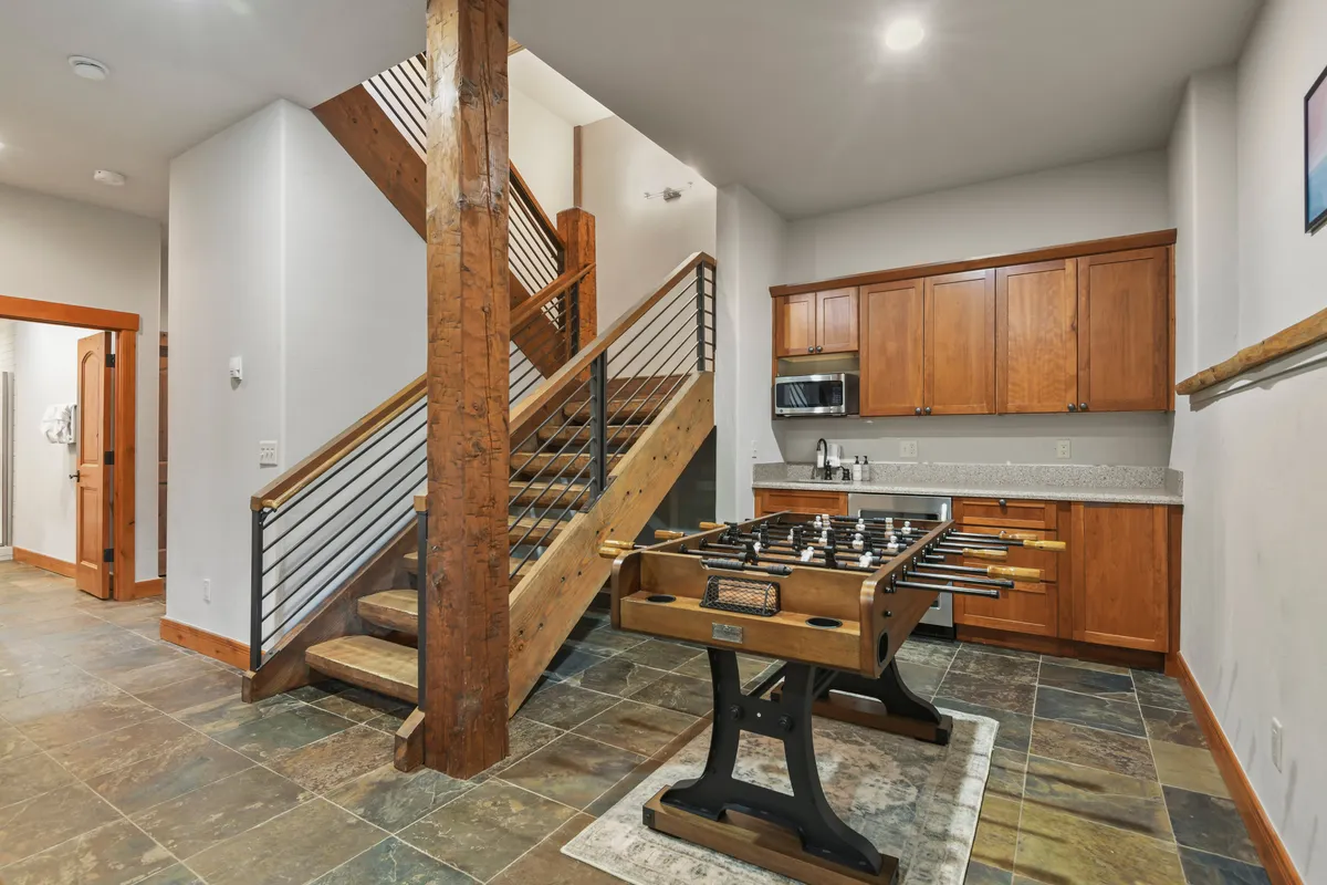 This image highlights the lower-level entertainment area featuring a foosball table, wooden staircase, and kitchen cabinetry. The playful setup invites friendly competition and socializing, while the adjacent kitchen space hints at easy access to the wet bar for mixing cocktails.