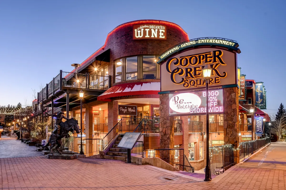 A vibrant outdoor plaza at Cooper Creek Square, featuring a prominent sign, a restaurant and wine shop entrance, ample outdoor seating, decorative lighting, and a horse sculpture. The space is lively and designed for communal experiences, with visible areas for shopping, dining, and enjoying live entertainment.