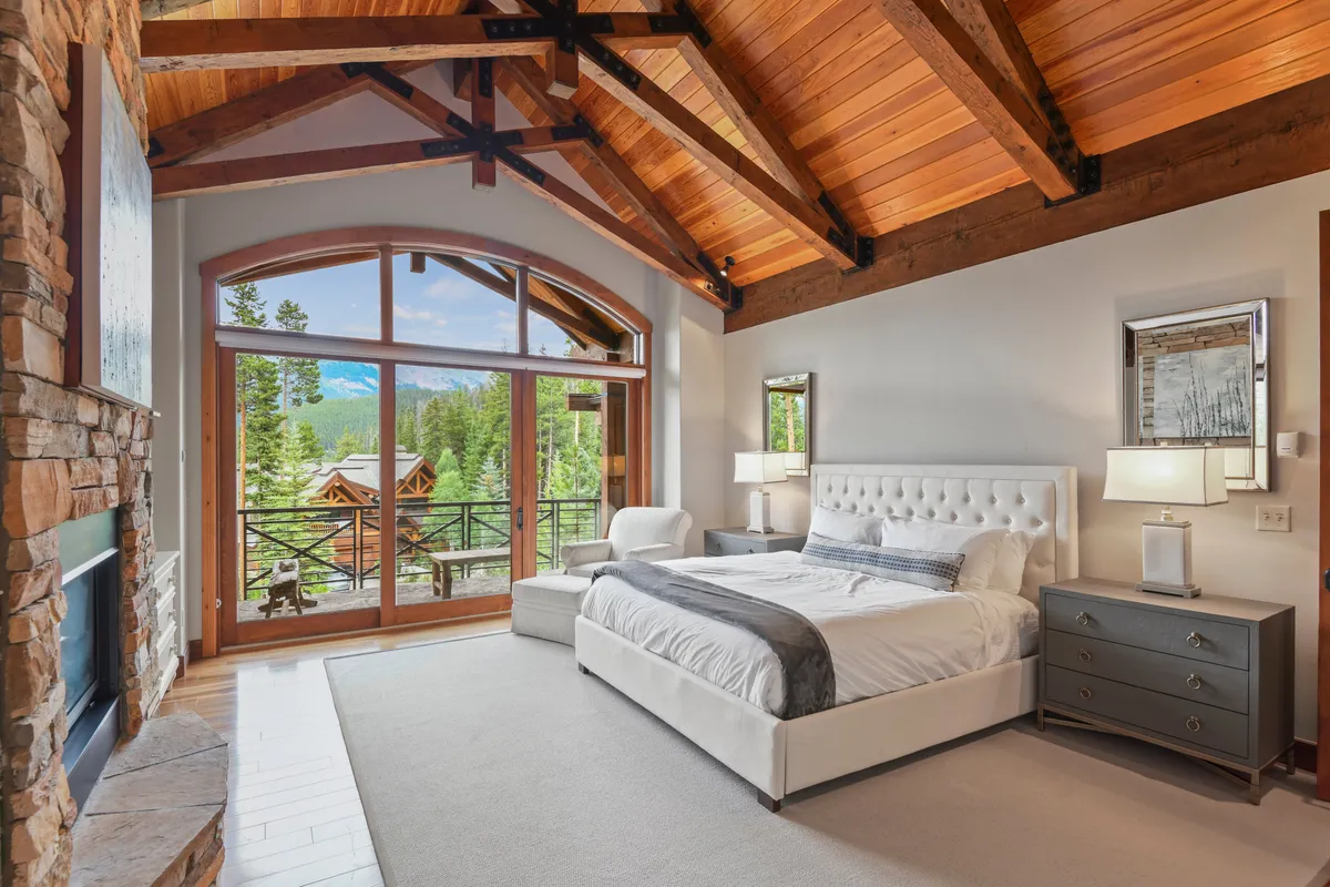 A spacious bedroom featuring a large bed with plush bedding, a cozy fireplace, and exposed wooden beams. Natural light pours in through the window, revealing a tranquil mountain view ideal for restful mornings or quiet evenings.