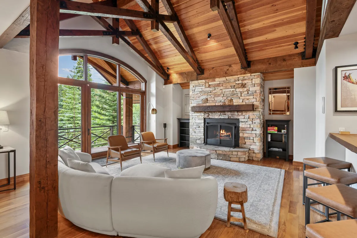 A sunlit great room featuring a stunning stone gas fireplace, vaulted ceilings with exposed beams, and a spacious arrangement of plush sofas. Towering windows reveal panoramic views of the Rocky Mountains, creating a seamless connection between the luxurious interior and breathtaking landscape.