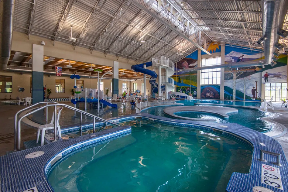 A dynamic indoor pool area at the Grand Park Rec Center, complete with water slides, a colorful mural, swimming pools, and lounge chairs. The environment is lively, family-friendly, and geared for fun activities for all ages.