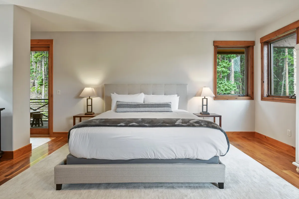 A serene bedroom featuring a king-sized bed, nightstands with warm lighting, and expansive windows framing a view of the outdoors. The room is styled for comfort and relaxation, inviting restful sleep or peaceful mornings with coffee.