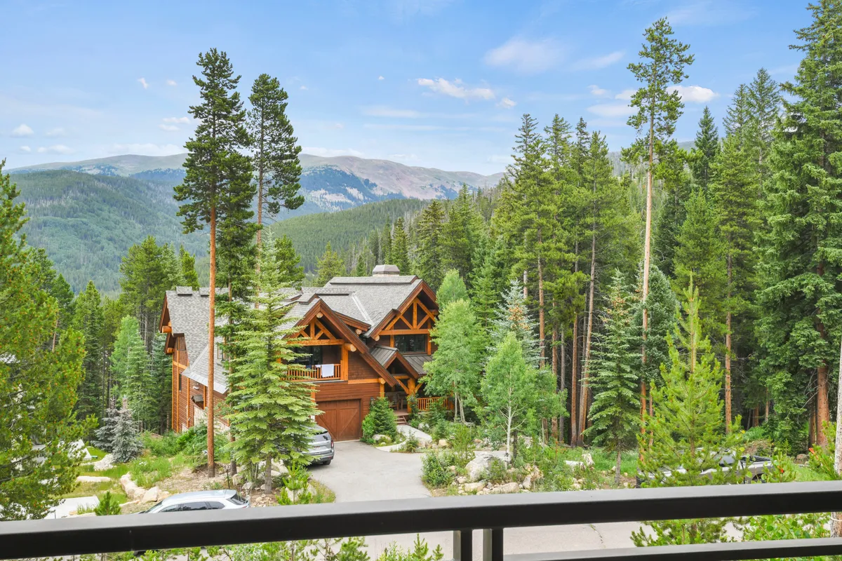 A stunning exterior shot of a luxurious log cabin set among tall pine trees, with a dramatic mountain backdrop. The scene emphasizes tranquility and a direct connection to the natural surroundings, inviting thoughts of morning coffee on the deck or evening stargazing.