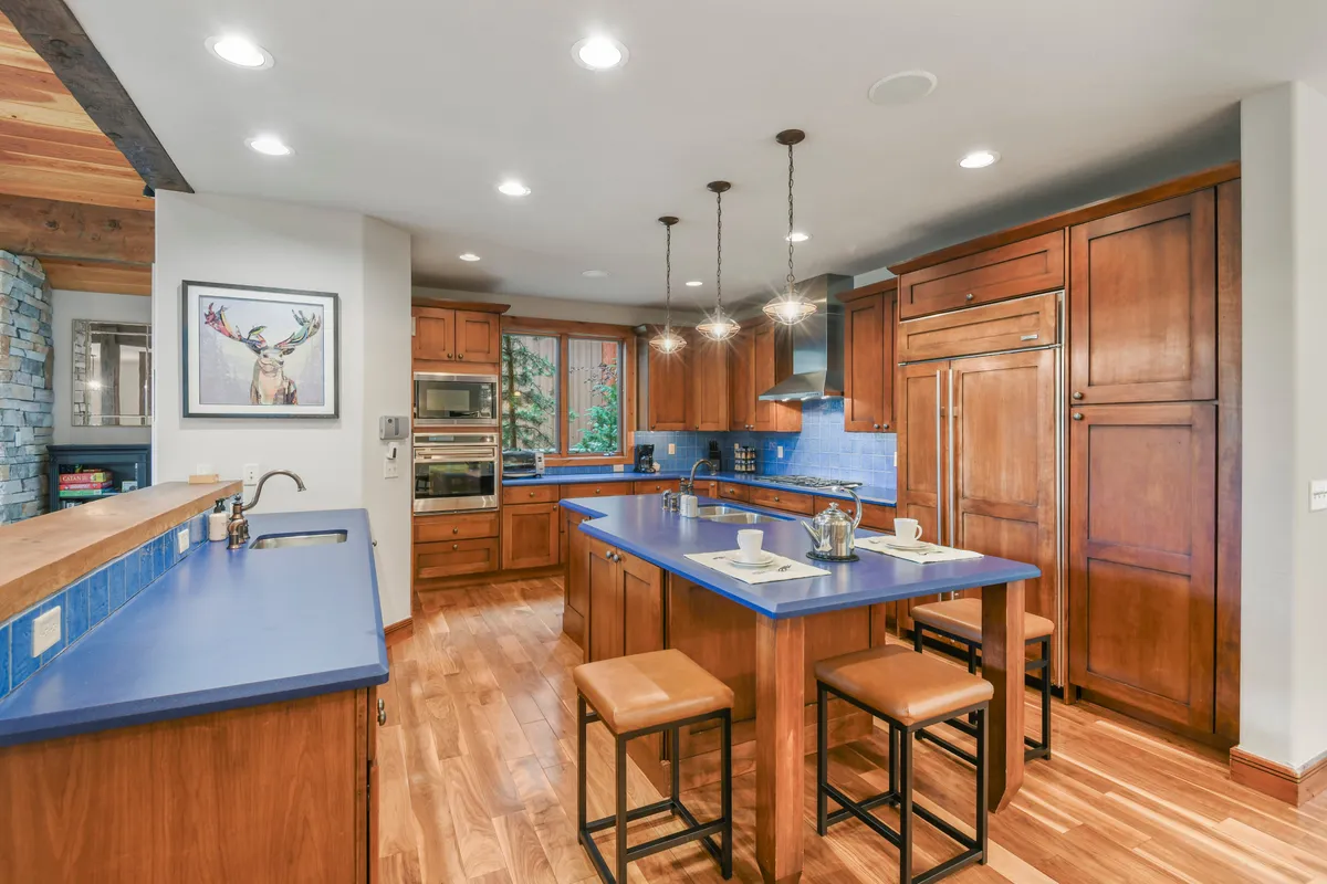 This image shows a spacious kitchen featuring a prominent blue countertop island, high-end wooden cabinetry, a refrigerator, and a stove. The layout is open and welcoming, with ample space for multiple people to cook or gather. The scene is lively and inviting, evoking the warmth of family baking sessions or group meal prep.