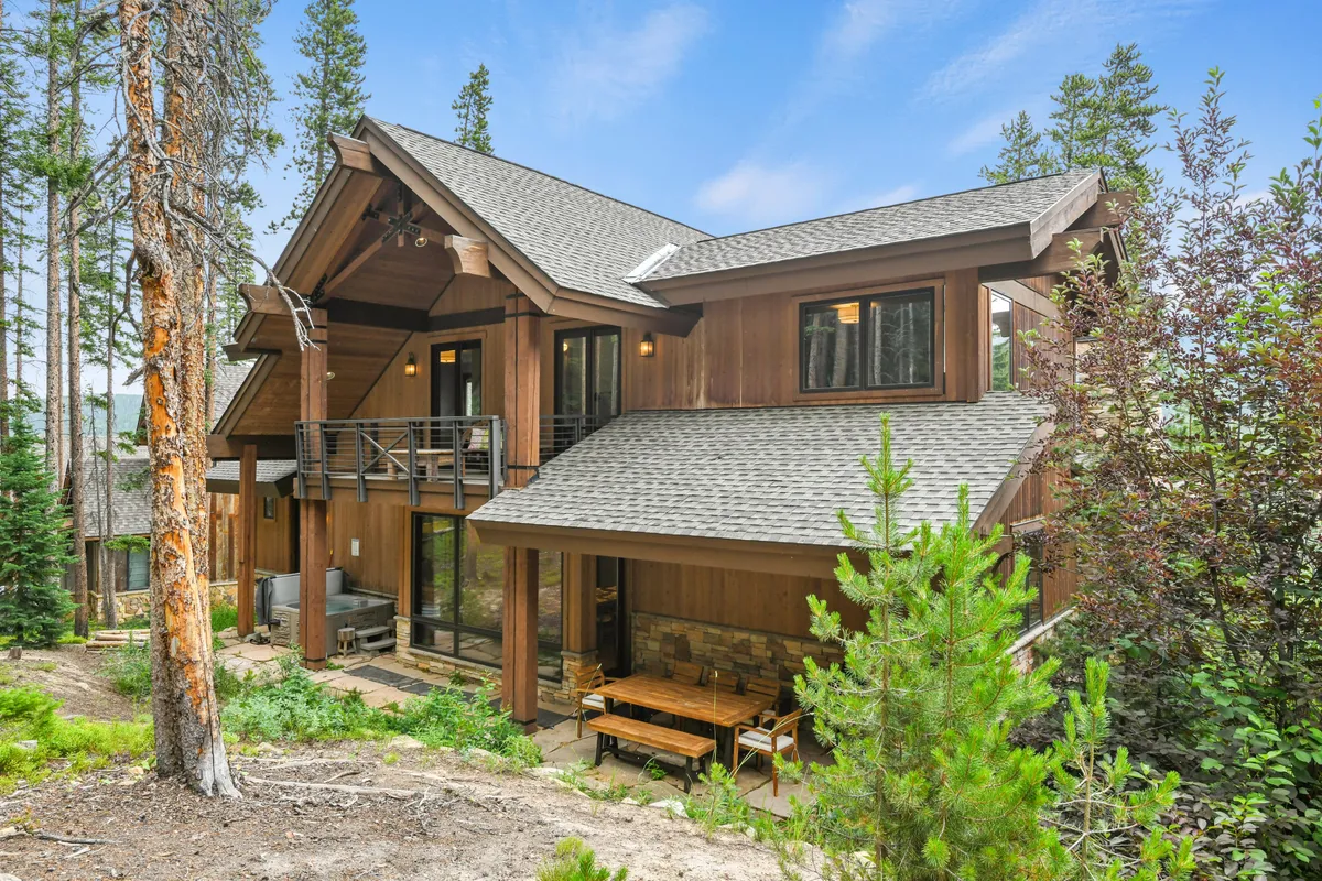 A wide exterior view of the chalet’s inviting wooden architecture, surrounded by lush trees. The outdoor dining area, visible on the patio, is set up for relaxed gatherings—a perfect setting for stargazing or enjoying the fresh mountain air.